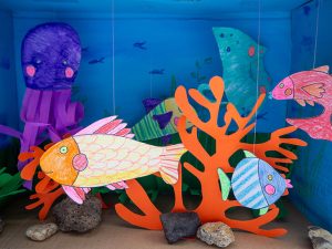 Nahaufnahme eines DIY-Aquariums aus einem Schuhkarton in dem bunt bemalte Fische aus Papier schwimmen.