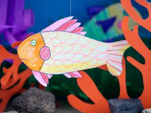 Ein großer, mit Buntstiften von STAEDTLER gestalteter Fisch aus Papier hängt in einem Aquarium aus einem Pappkarton.