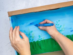 Jemand malt kleine dunkelblaue Fische mit einem Wachsmalstift von STAEDTLER an die Rückwand eines DIY-Aquariums aus Pappe. Foto: Johanna Rundel