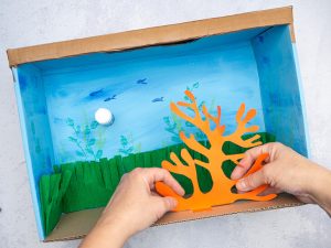 Jemand klebt eine Koralle aus Fotokarton in ein DIY-Aquarium aus Pappe