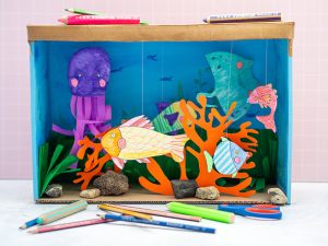 Ein selbstgebasteltes Aquarium aus einem Pappkarton steht vor einer rosa Wand. Davor und darauf liegen Stifte von STAEDTLER. 
