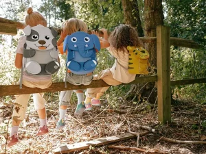 Drei Kinder sitzen mit Tier-Rucksäcken von Affenzahn auf einem Holzzaun.