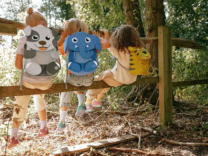 Drei Kinder sitzen mit Tier-Rucksäcken von Affenzahn auf einem Holzzaun.