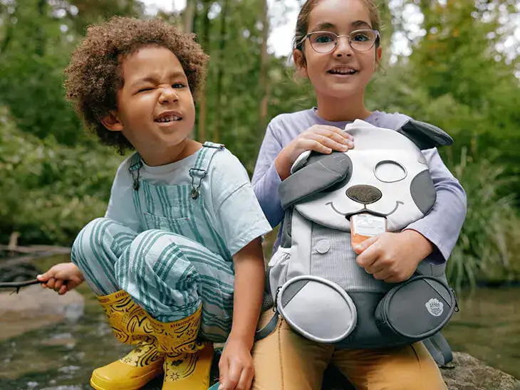 Zwei Kinder hocken mit einem Kindergartenrucksack von Affenzahn im Grünen.