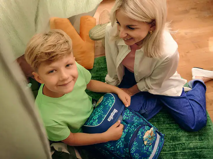 Ein Junge sitzt mit seiner Mutter und seinem Schulranzen auf dem Boden.